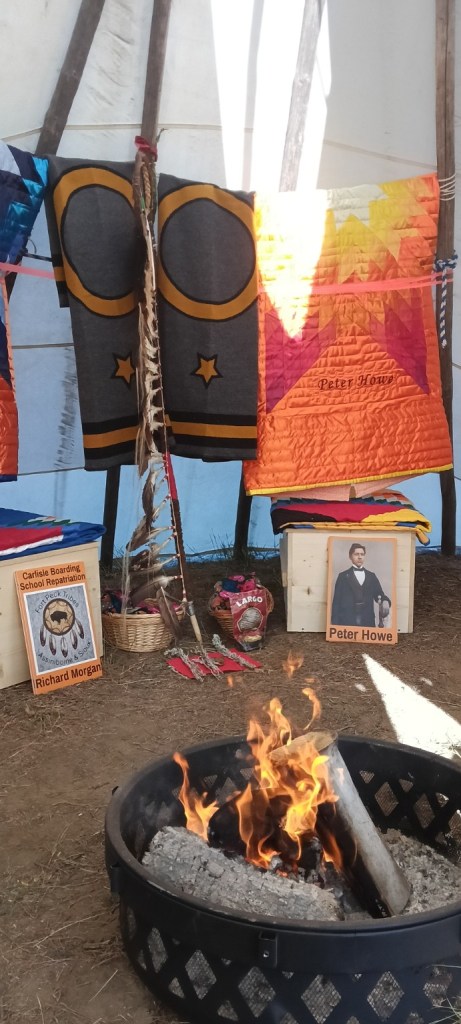 A fire burns inside the tipi where Peter Howe and Richard Morgan were kept before burial, with star quilts, photographs, eagle feathers and memorial items arranged behind it.