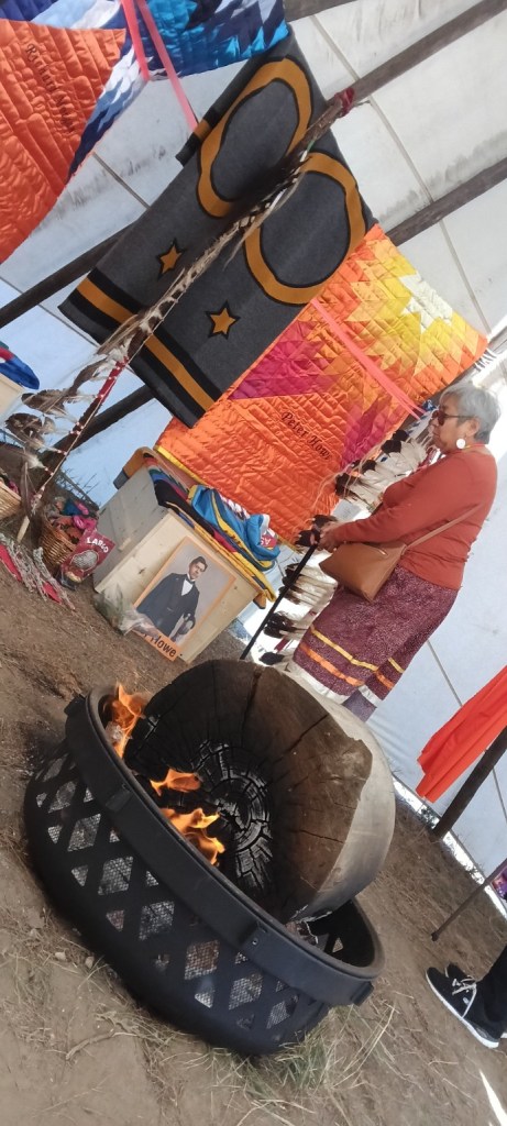 A woman stands quietly inside the tipi near Peter Howe’s memorial display while a fire burns in the foreground beneath hanging star quilts and eagle feathers.