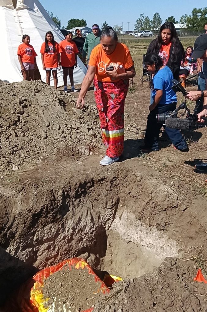 A Fort Peck community member drops a handful of earth into one of the graves of Peter Howe and Richard Morgan during their burial, while people in orange shirts stand nearby.