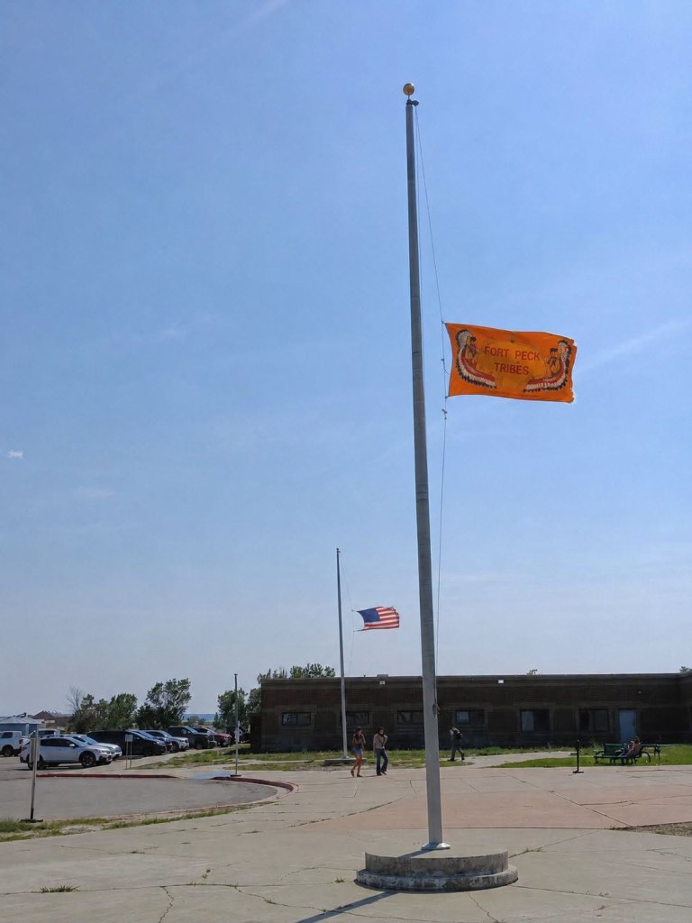 Orange Fort Peck Tribal Flag and U.S. flag fly at half-staff in Poplar following the deaths of former Tribal Chairman A.T. “Rusty” Stafne and Dolly Parton.