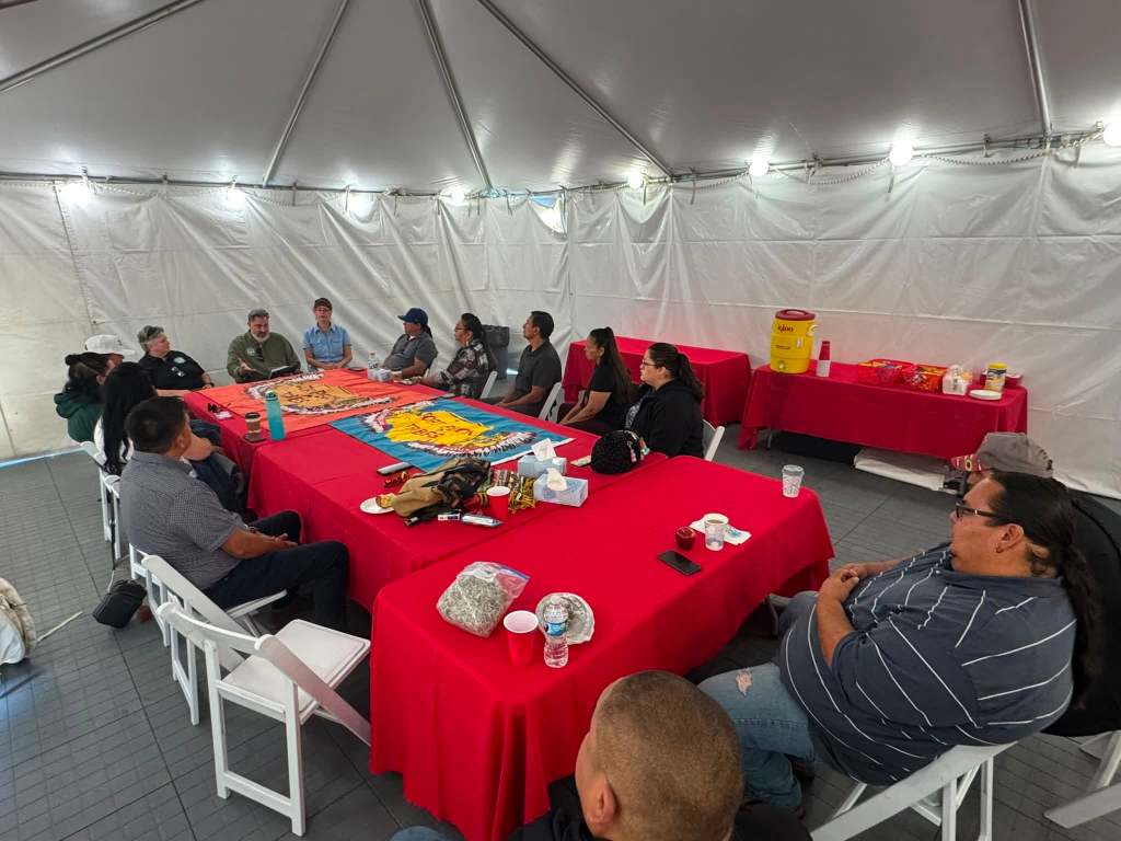 Fort Peck delegation seated around tables during an orientation meeting at Carlisle, with blue and orange Fort Peck Tribal flags displayed on the table.