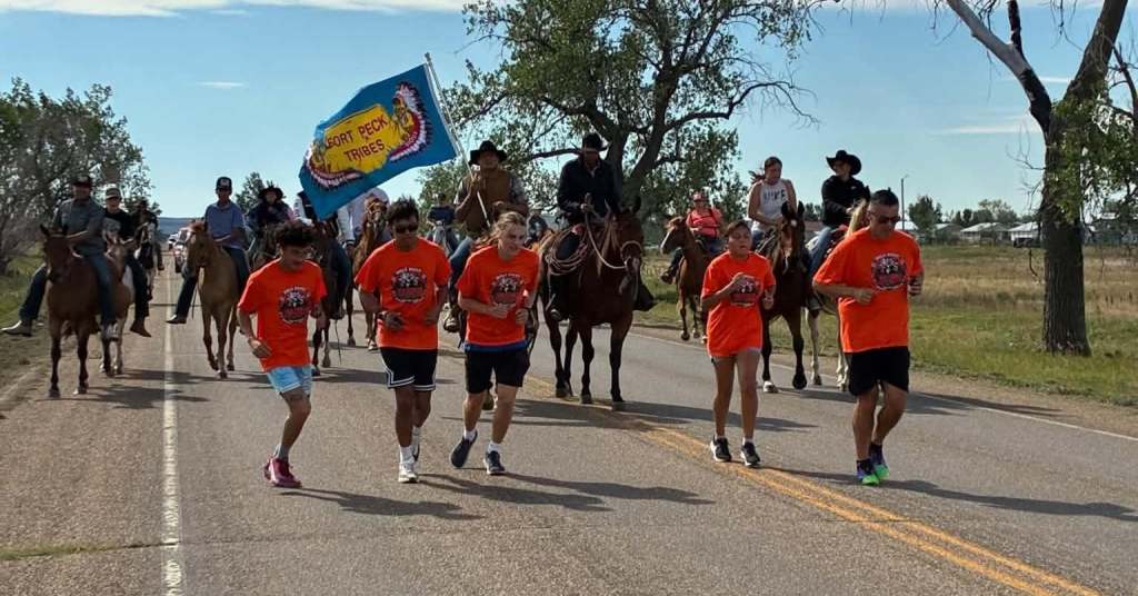 Wolf Point Wolves Cross Country runners wearing orange shirts run along a rural road while horseback riders follow behind, including a rider carrying the Fort Peck Tribes flag, during the welcome-home procession for Peter Howe and Richard Morgan.