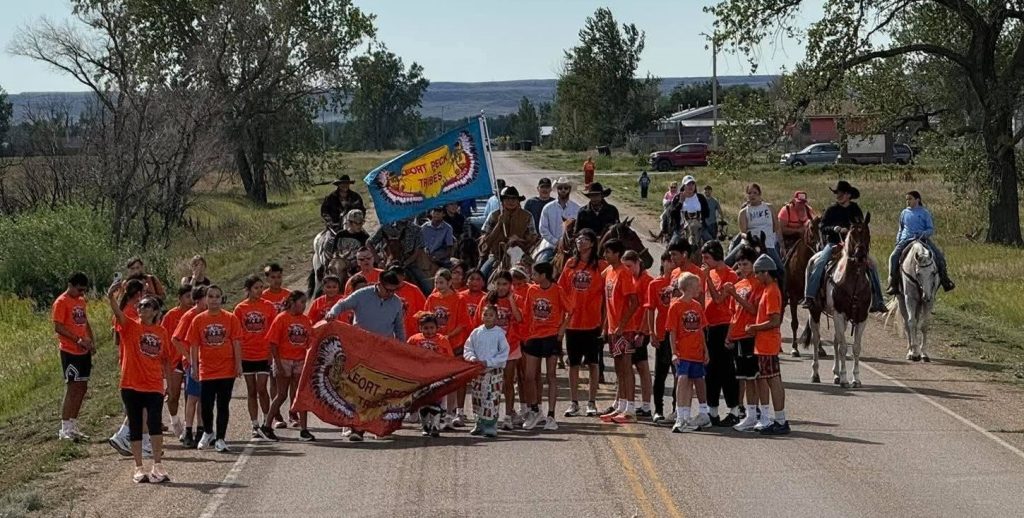 A large group of students and community members wearing orange shirts gathers on a rural road with horseback riders behind them carrying Fort Peck Tribal flags during the Carlisle boarding school reburial procession.