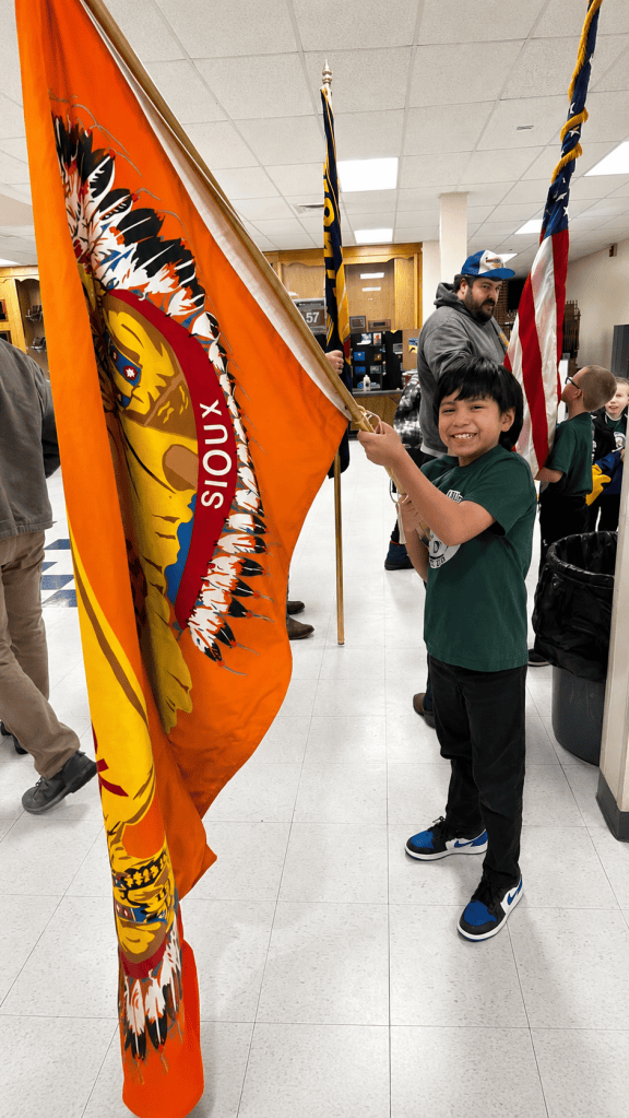 Smiling young student holds an orange Fort Peck Tribal Flag inside a school.