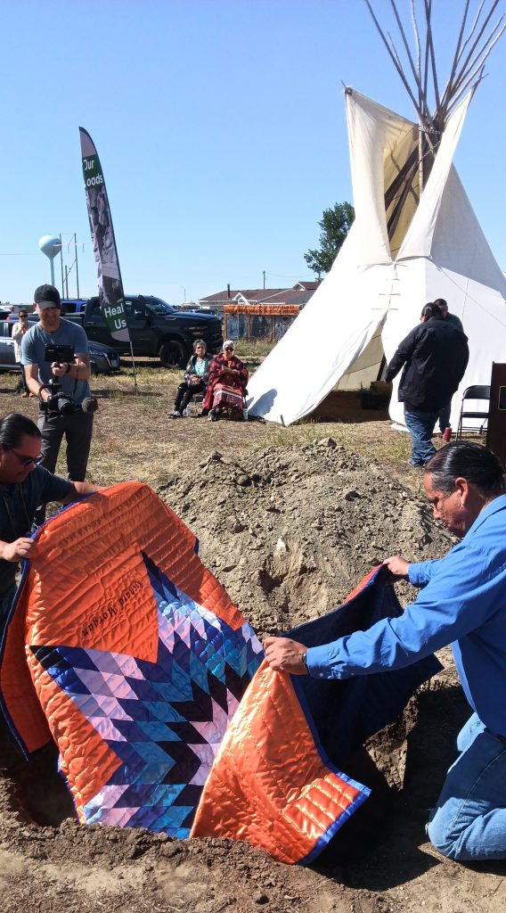 Two men arrange an orange star quilt around a burial box beside an open grave, with a tipi standing behind them.