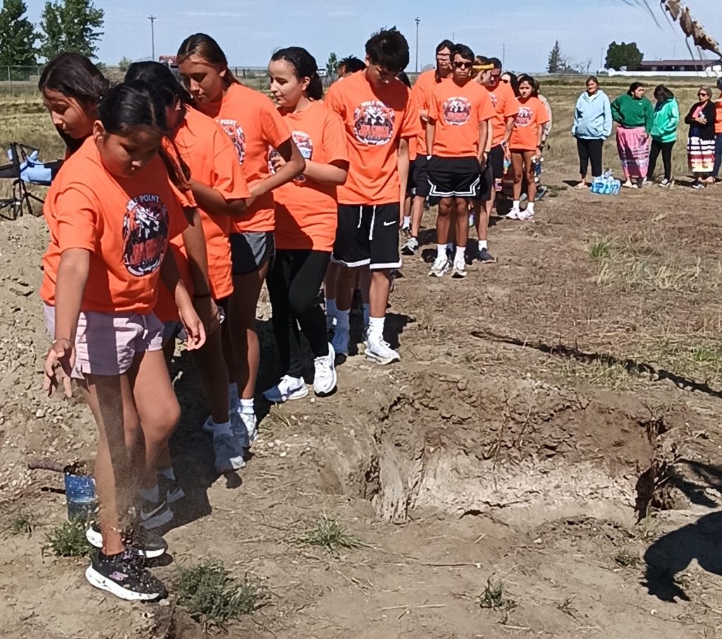 Youth wearing orange shirts file past the open graves of Peter Howe and Richard Morgan, placing handfuls of earth into the graves as an eagle staff is held nearby.