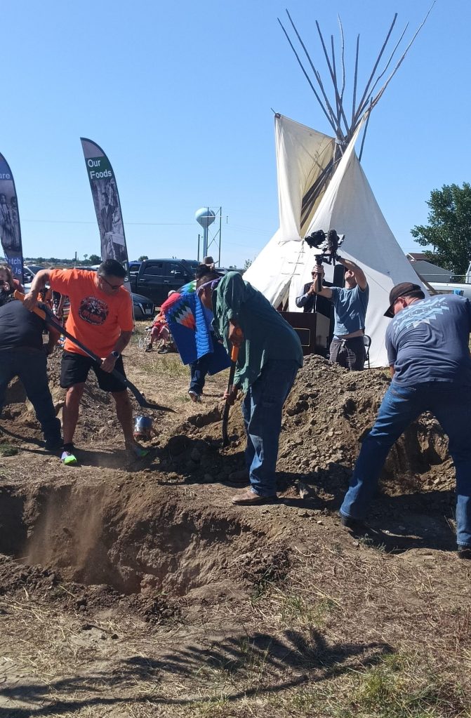 Men use shovels to fill the graves of Peter Howe and Richard Morgan by hand following their burial ceremony, with a tipi in the background.