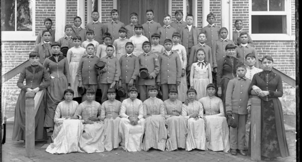 Historical black-and-white group photograph of Fort Peck Assiniboine and Sioux children posed in rows outside a brick building at the Carlisle Indian Industrial School in Pennsylvania.